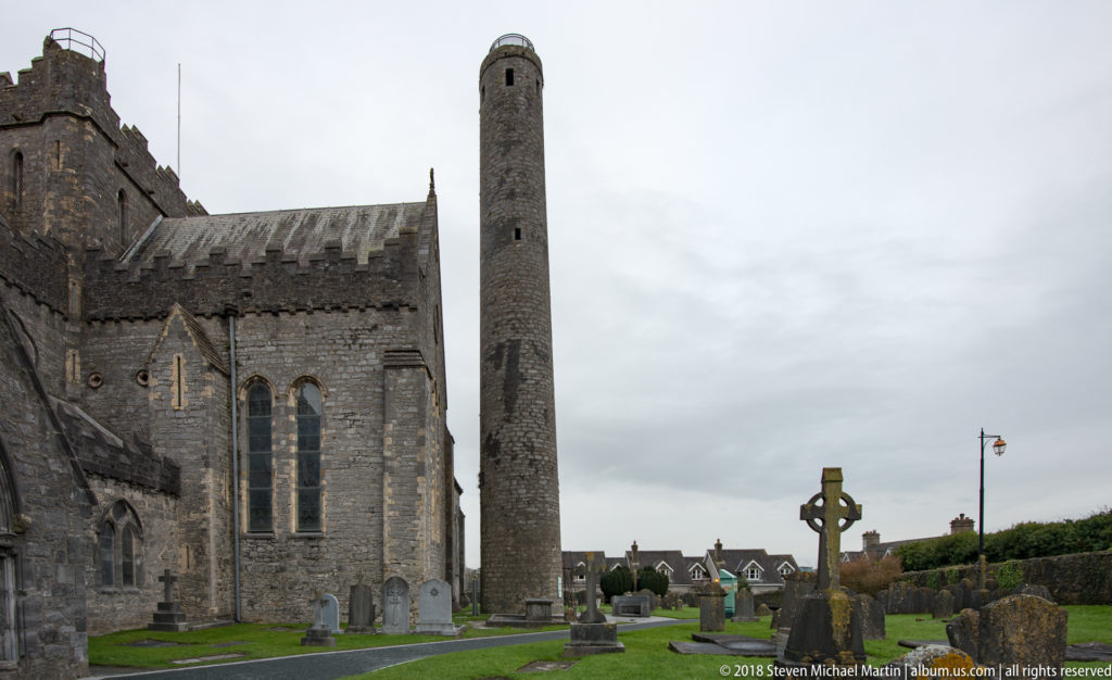 Stone cathedral wall and round tower rise behind a Celtic-style graveyard cross and clustered memorials.