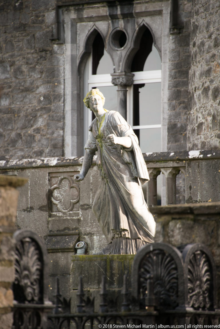 Weathered stone sculpture of a robed figure before a Gothic window and carved stone parapet.