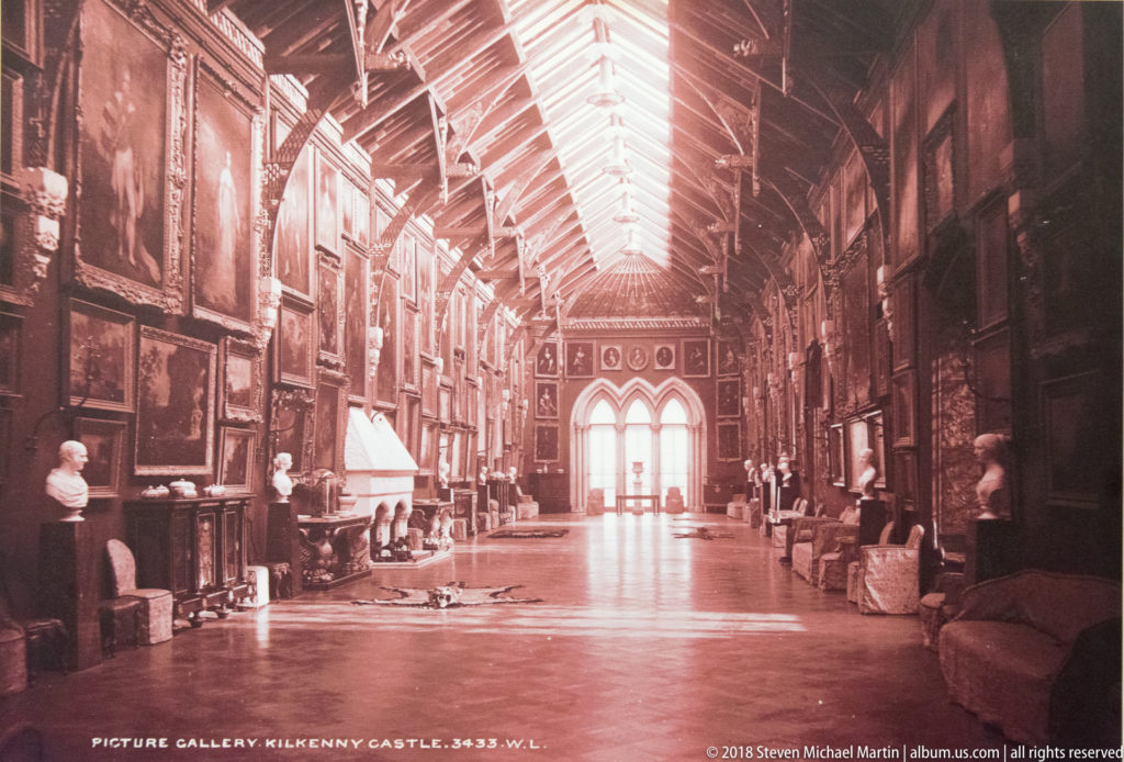 Historic monochrome view labeled Picture Gallery, Kilkenny Castle, showing densely hung paintings, furniture, glazed roof, and pointed end window.