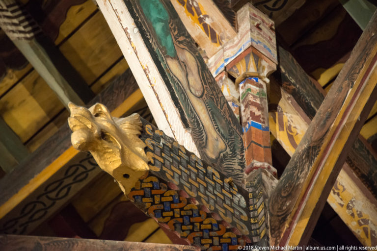 Close view of a gilded carved head at the junction of painted timber beams decorated with interlace and geometric patterns.