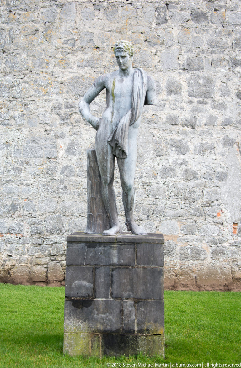 Bronze figure sculpture leaning forward on a pedestal beside a rough stone wall and closely cut green lawn.