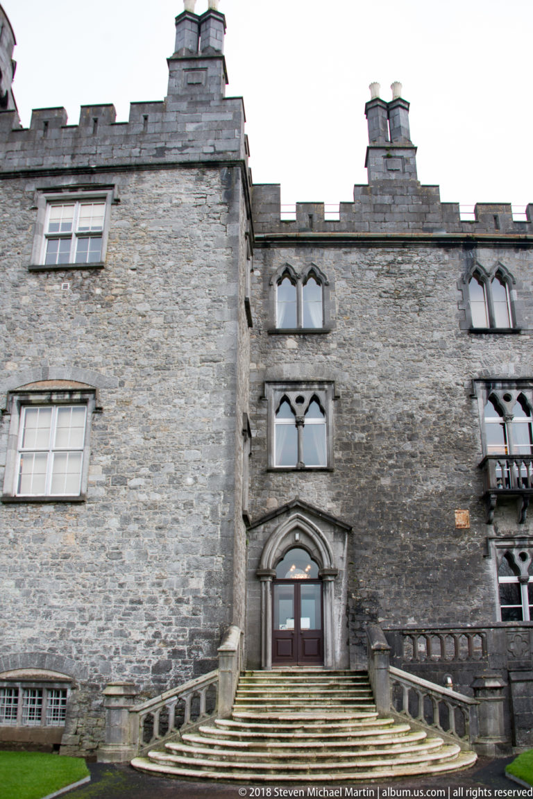 Grey stone building with steps to a pointed doorway, multi-light windows, chimneys, and a crenellated parapet.
