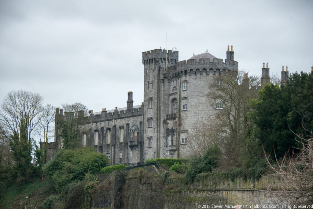 Kilkenny Castle above trees and a stone embankment, dominated by a large round crenellated tower and long gray façade.