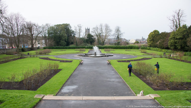 Formal garden with central path, fountain, clipped lawns, low planting beds, two visitors, and trees in the distance.
