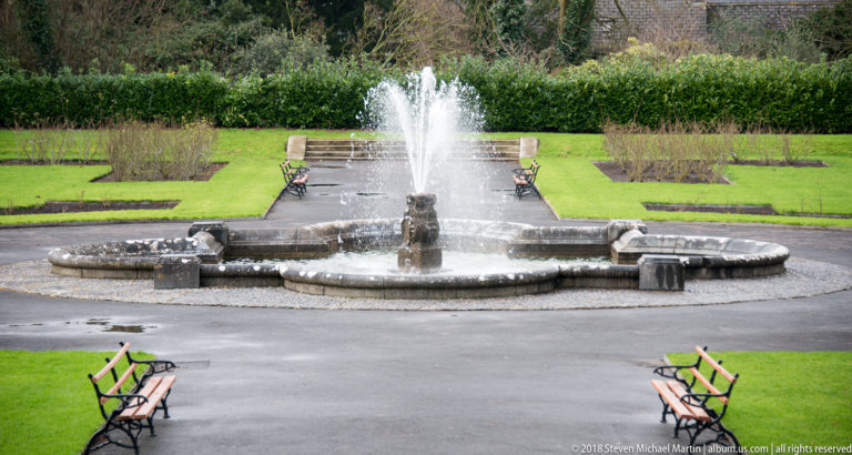 Circular stone fountain spraying water in a formal garden, with low beds, hedges, paths, and benches arranged symmetrically.