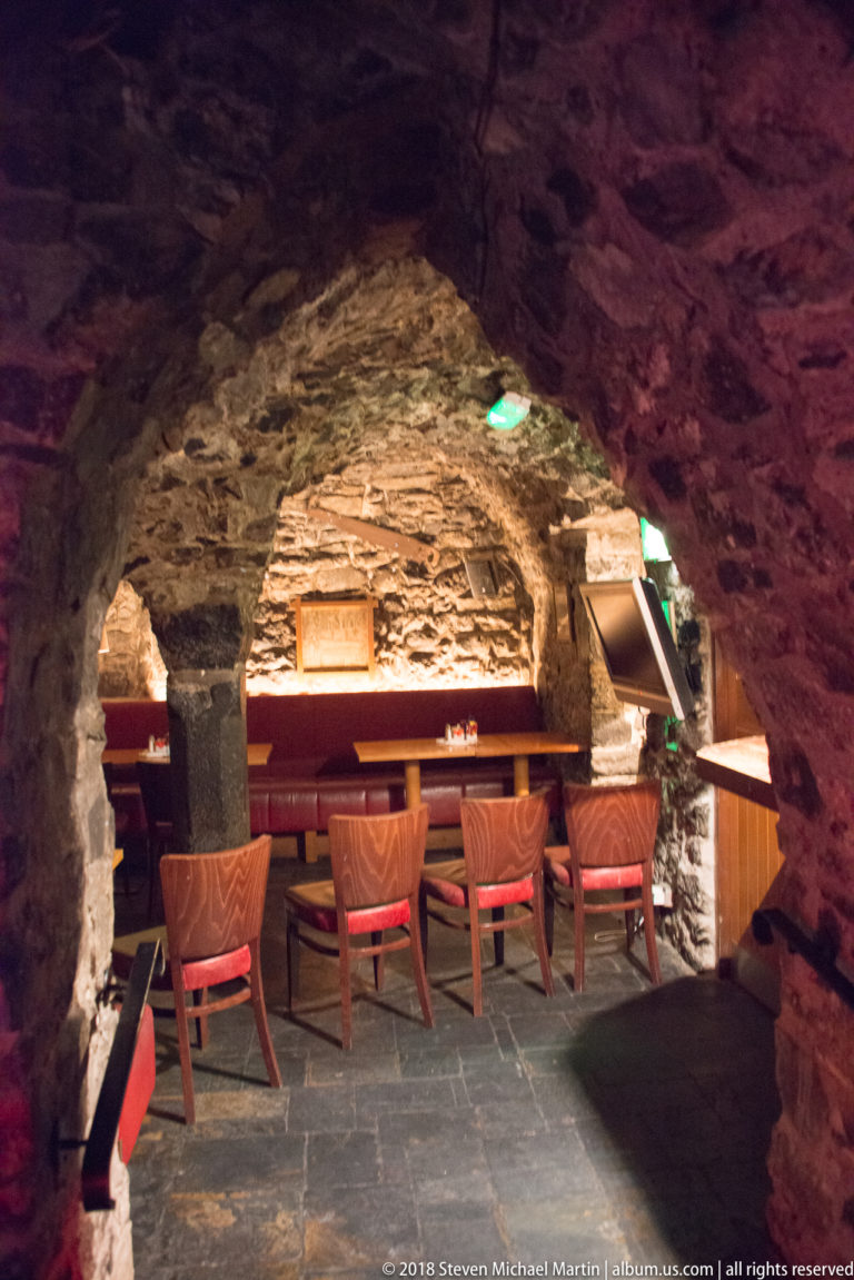 Low stone-vaulted alcove with upholstered chairs, narrow tables, warm lighting, and slate floor inside a historic pub.