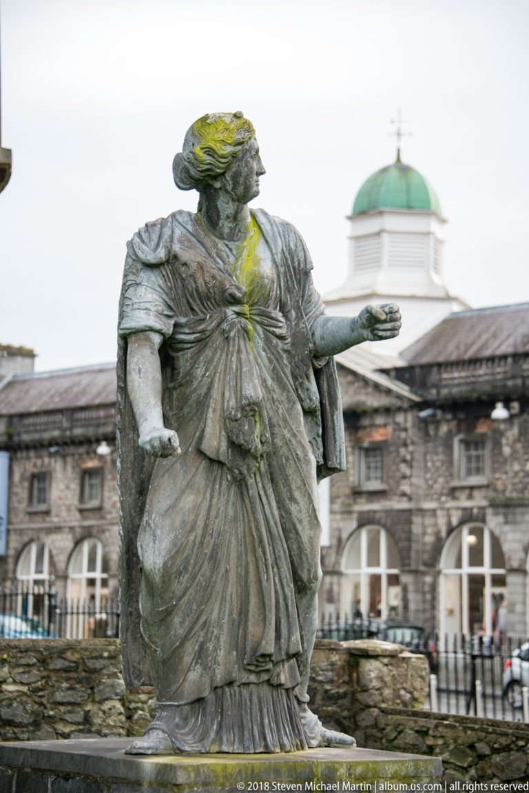 Weathered bronze draped female figure with one arm extended, set before stone buildings and a pale cupola.