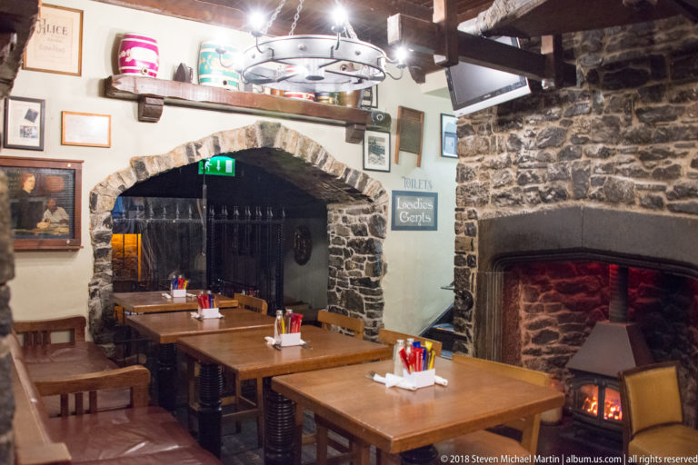 Wooden tables beneath stone arches beside a glowing fireplace in a rustic pub dining room.
