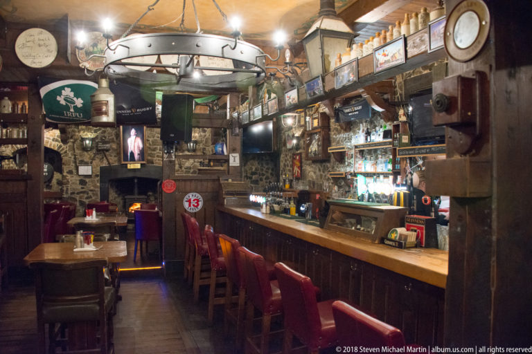 Long wooden pub bar with red stools, exposed stone walls, taps, clocks, framed memorabilia, and television screens.