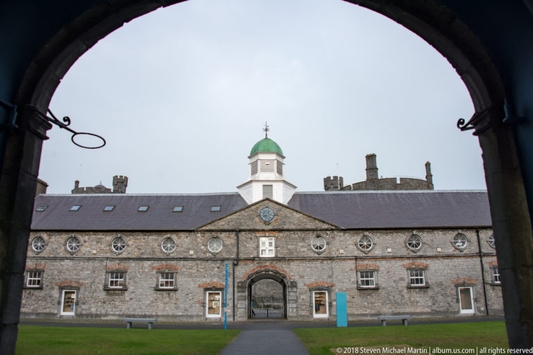 Stone courtyard building framed by a dark arch, with central entrance, clock, circular upper windows, and white cupola with green roof.