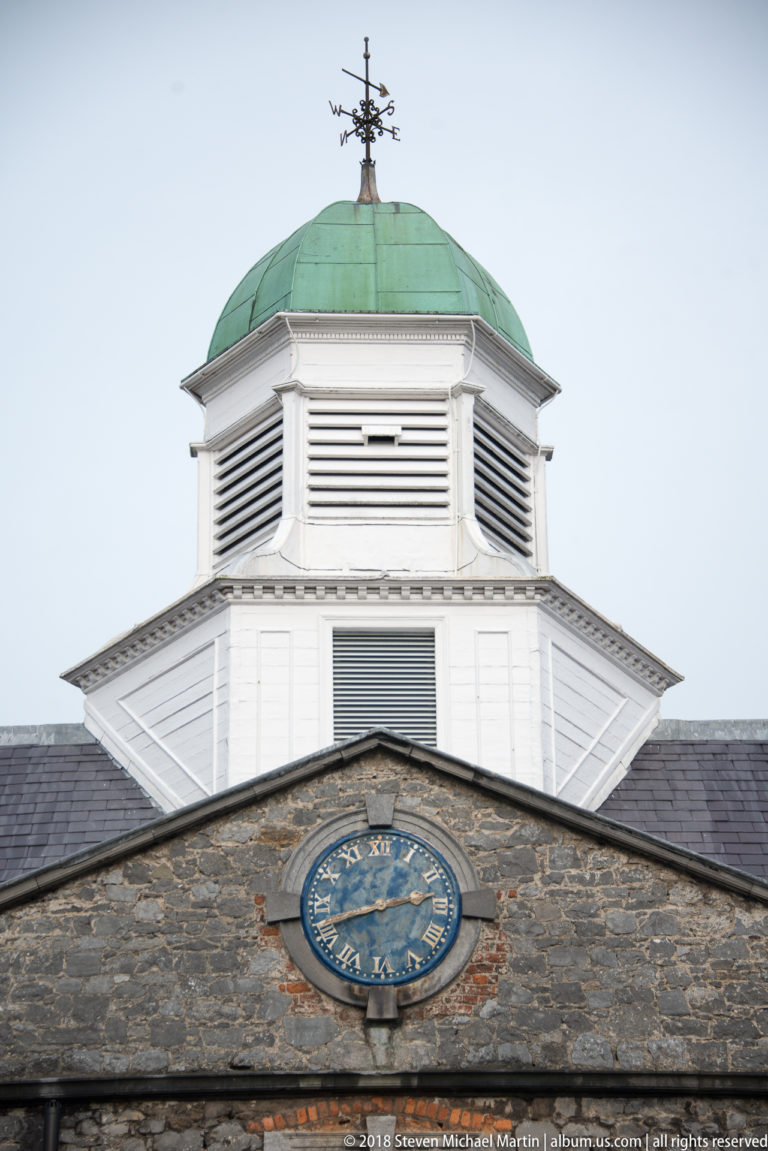 White louvered cupola with green metal roof and weather vane above a rough stone gable with a blue clock face.