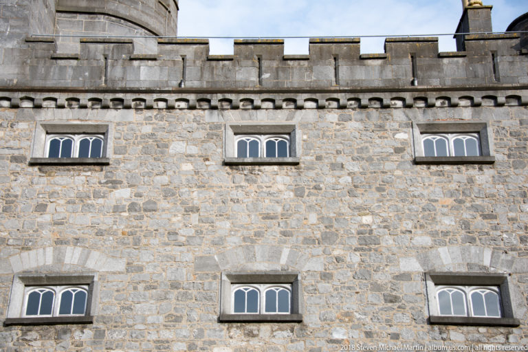 Limestone castle wall with two rows of small multi-light windows beneath a crenellated parapet.