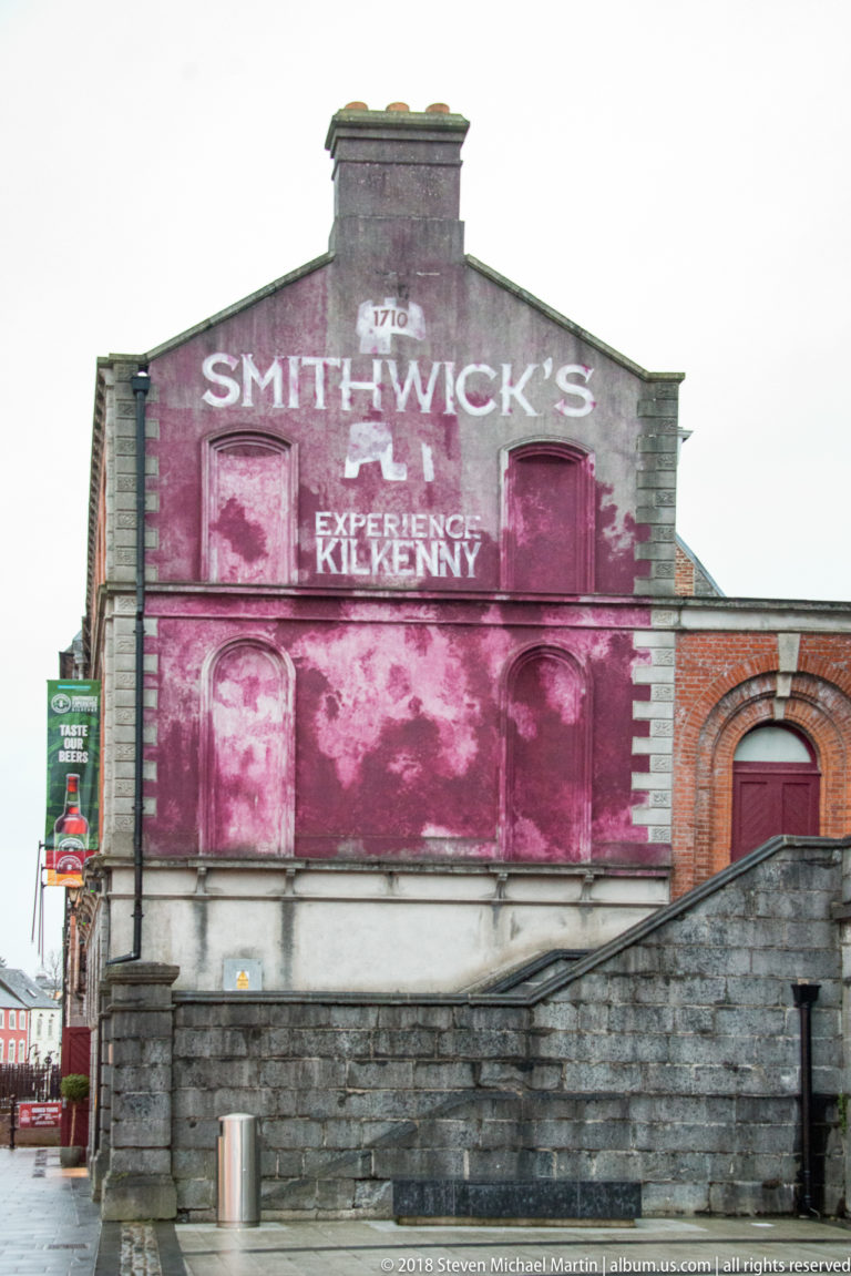 Stone and brick building with a large faded magenta sign reading Smithwick’s Experience Kilkenny across the upper façade.