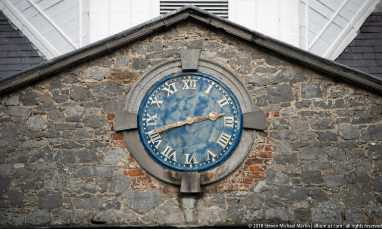 Blue clock with gold hands and Roman numerals set into rough grey stone masonry with dressed stone surround and traces of red brick.