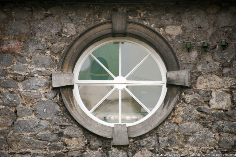 Round window with white radial muntins and a dressed stone surround set into rough grey masonry.