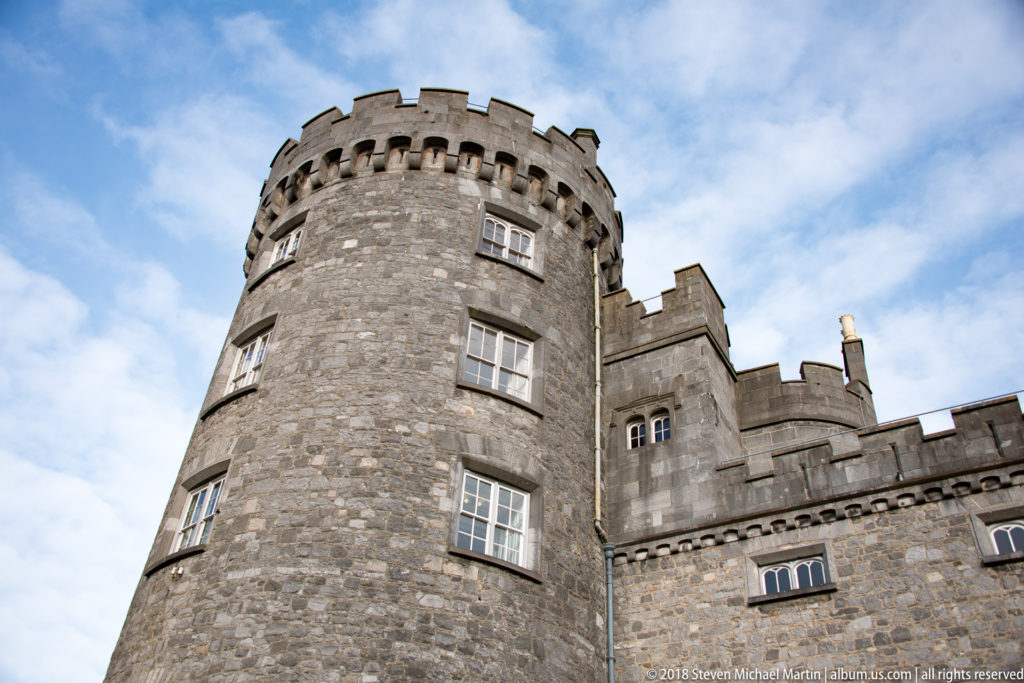 Round stone tower of Kilkenny Castle rising above adjoining walls beneath a partly cloudy sky.