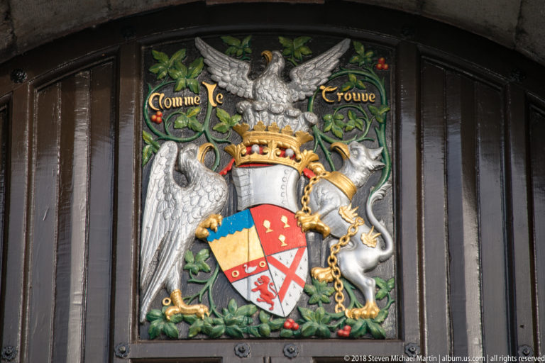 Painted coat of arms with crowned helm, shield, animal supporters, foliage, and the motto “Comme je trouve.”