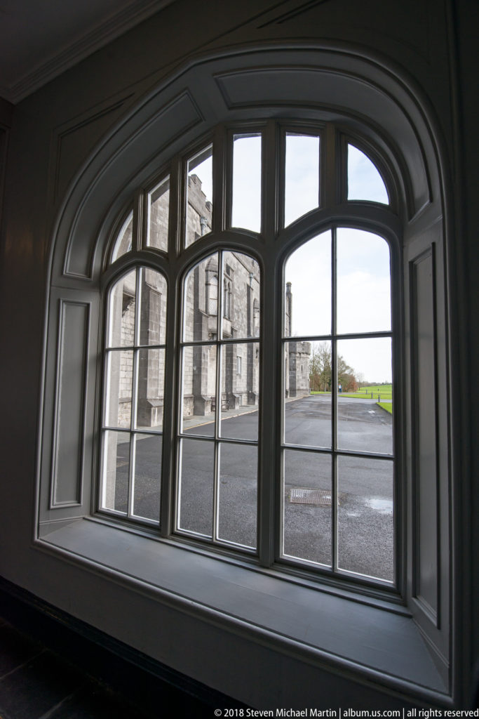 Large arched multi-pane window looking from the castle interior toward stone buildings, pavement, and lawn.