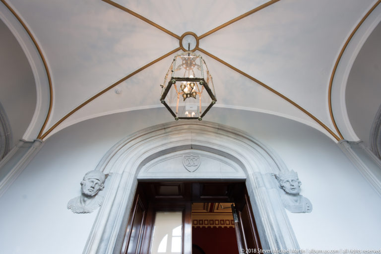White vaulted passage with hanging lantern, arched doorway, and sculpted stone heads on both sides.