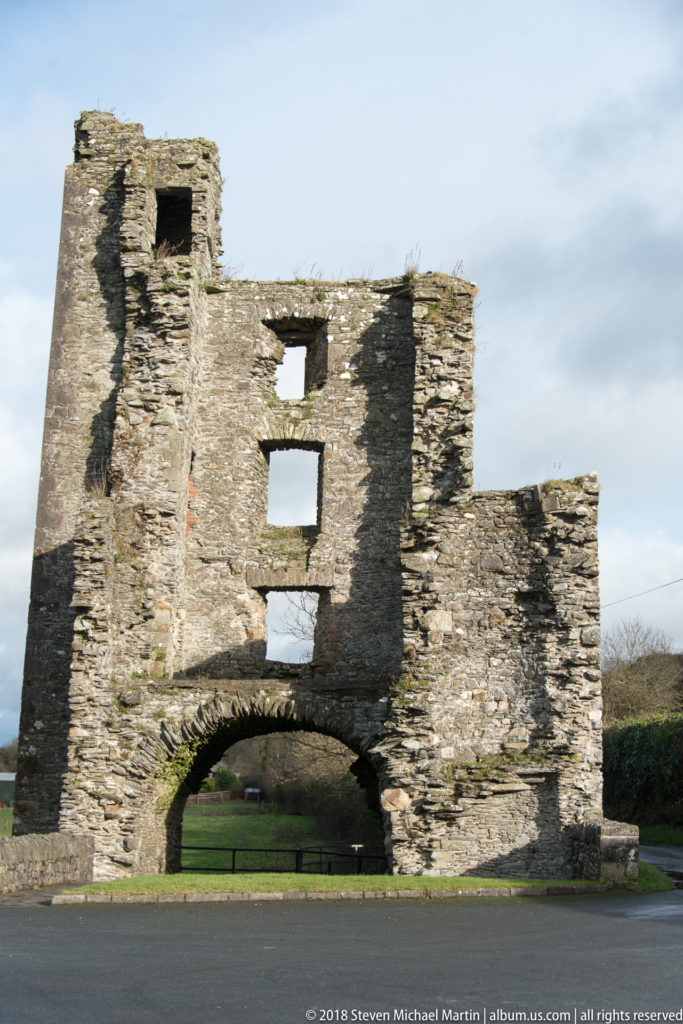 Roofless stone gateway ruin with a broad round arch, rectangular upper openings, and grass visible through the passage.