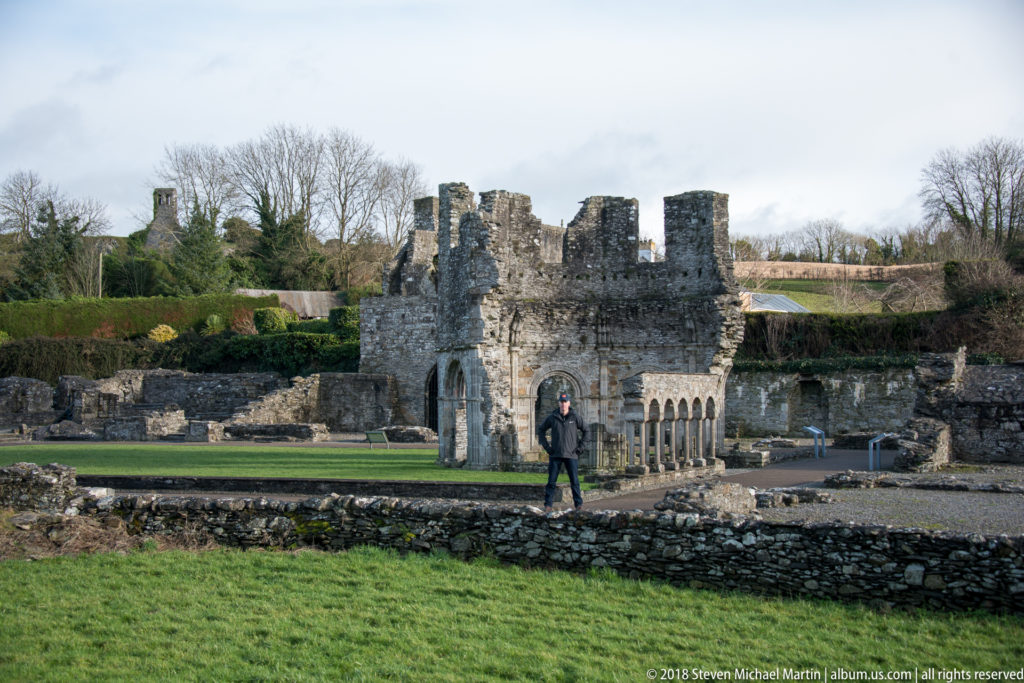 Ruined lavabo with arched openings stands beyond low stone foundations, with one visitor providing scale.