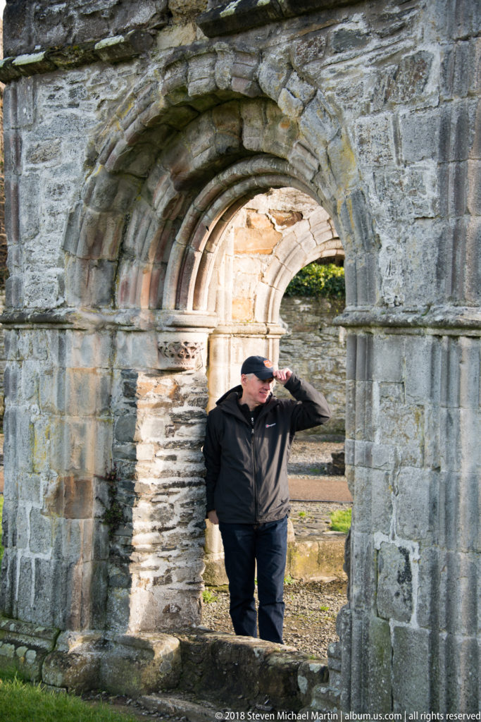 Person in dark clothing stands beneath a thick recessed stone arch with sunlit ruins visible beyond.
