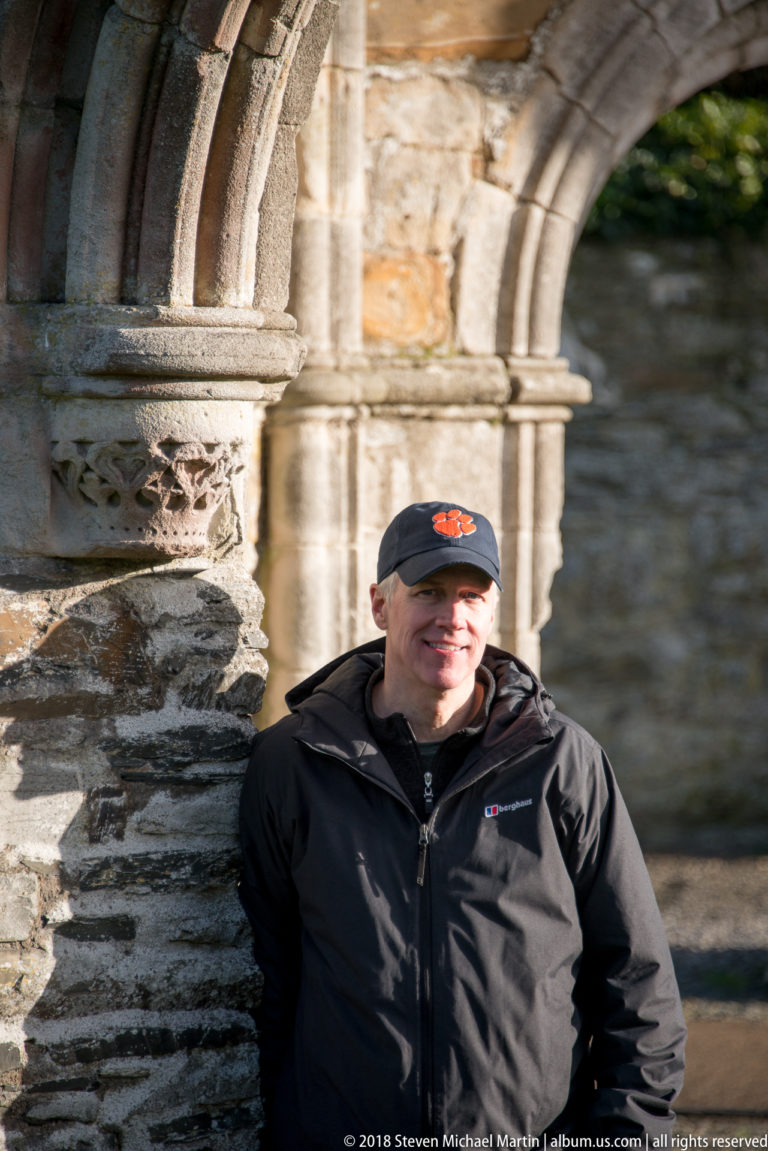 Smiling visitor in a dark jacket and cap leans beside a sunlit carved stone arch and rough masonry wall.