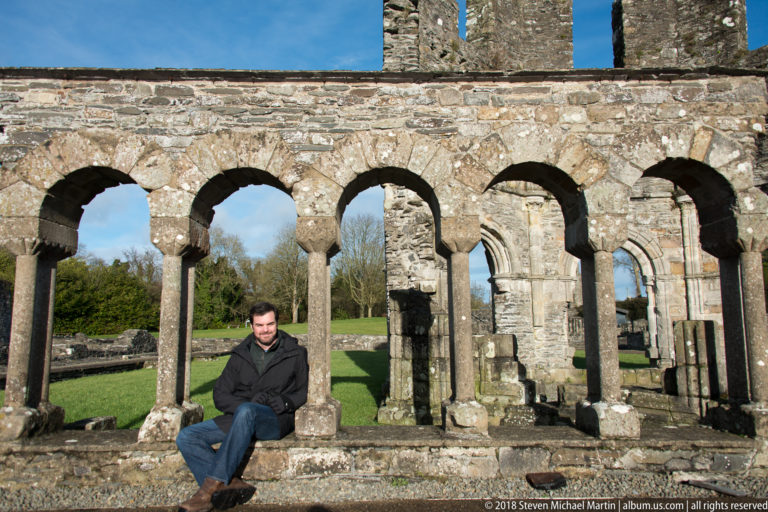 Seated visitor beneath a row of small round stone arches and slender columns, with green lawn visible through the arcade.