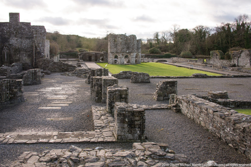 Elevated view across low stone foundations, gravel paths, lawns, and several standing sections of the ruined abbey.