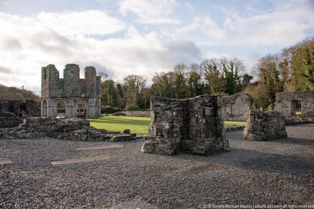 Low stone foundations and gravel paths lead toward taller roofless abbey walls beside a rectangular lawn.