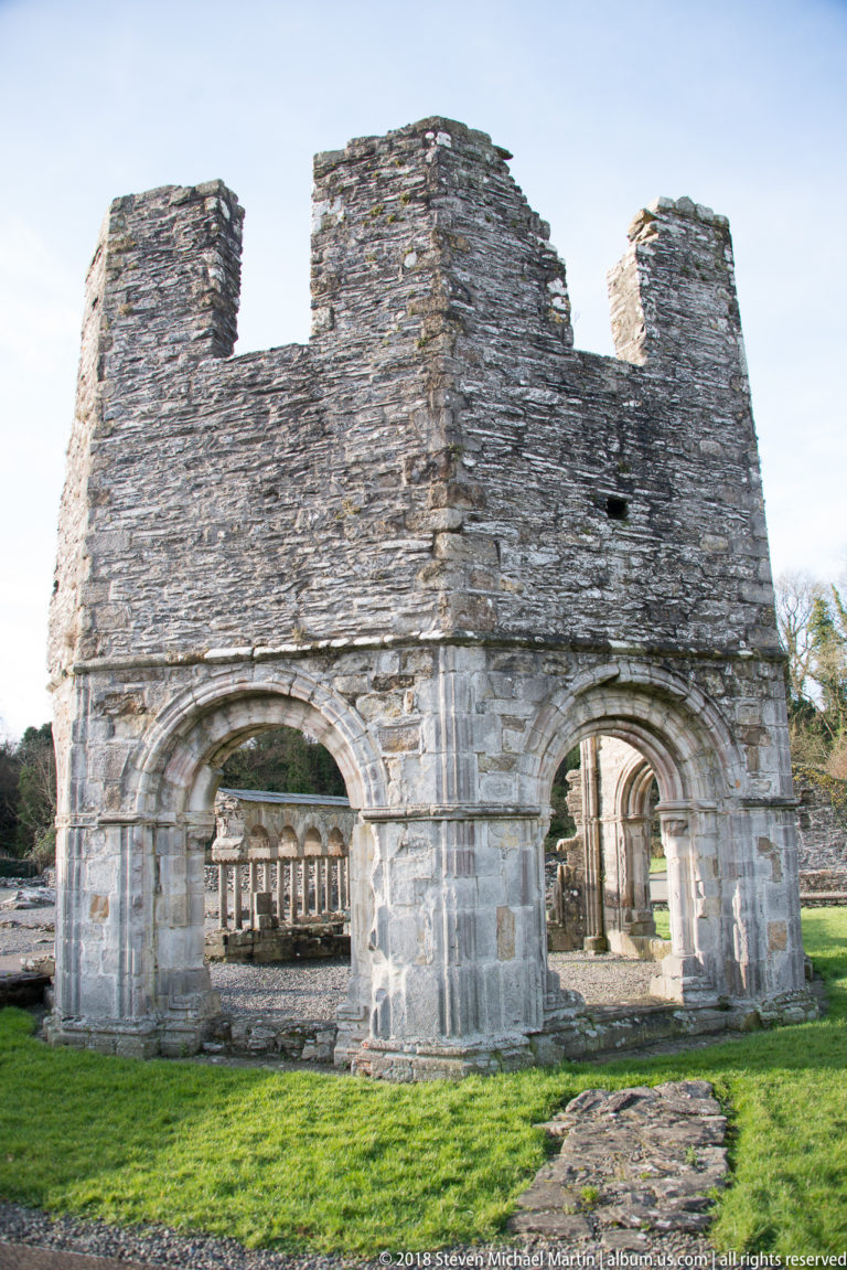 Ruined polygonal stone lavabo with paired round arches, heavy piers, and a broken masonry wall above.
