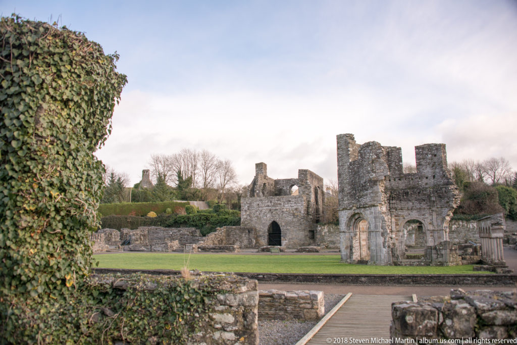 Wide view across lawns and low walls toward roofless abbey ruins, framed at left by ivy-covered masonry.