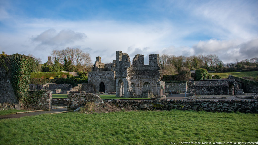 Green foreground slopes toward clustered roofless abbey ruins, low stone walls, hedges, and leafless trees.