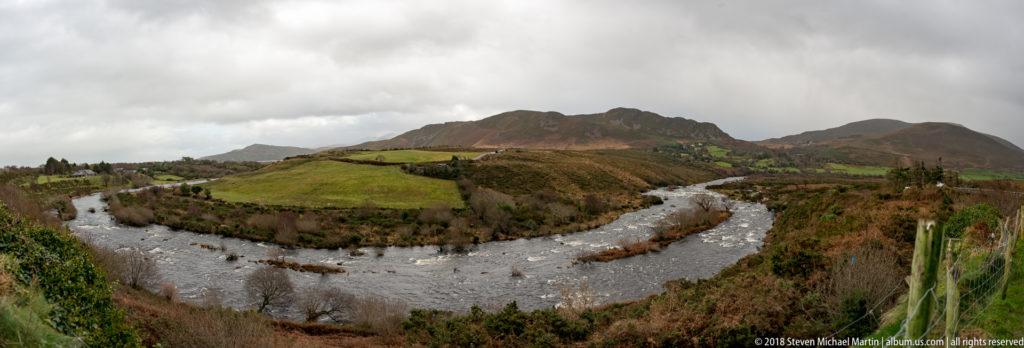 Fast-moving river bends around green pasture beneath rugged, cloud-covered hills in the Ring of Kerry.