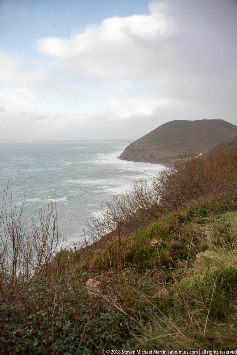 Rounded coastal headland beyond scrubby grassland and a pale blue-gray bay under clearing clouds.