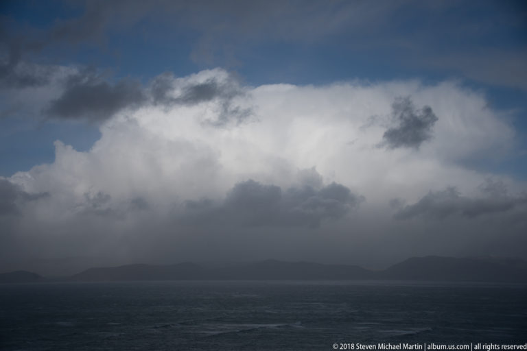 Large white and dark storm clouds rise above a nearly black sea and faint mountain horizon.
