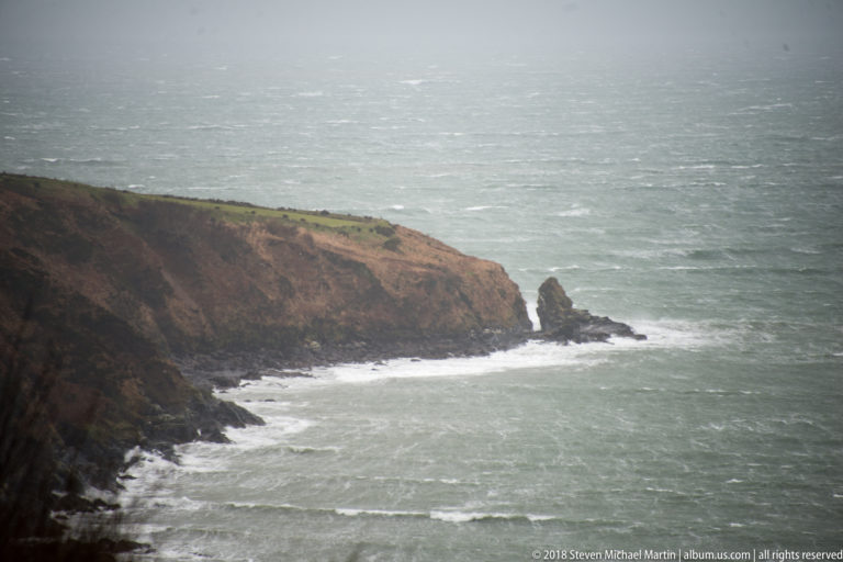 Brown rocky headland with a narrow offshore sea stack surrounded by white surf.