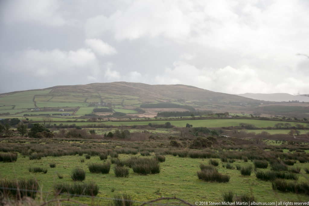 Green fields divided by hedges and walls across rolling hills, with rough rush-filled pasture in front.