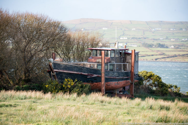 Weathered wooden fishing boat hauled onto grass beside trees, with an inlet and fields in the background.