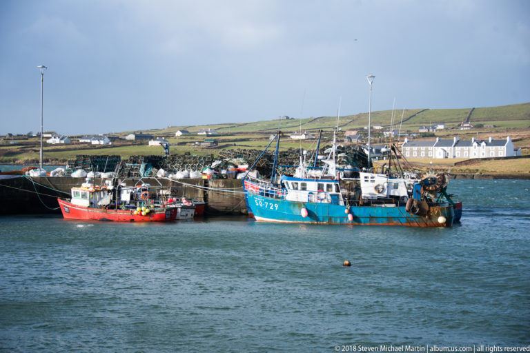 Several brightly colored commercial fishing boats moored together in a sheltered harbor below green fields and white houses.