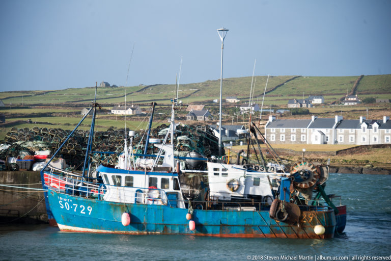 Blue commercial fishing boat moored at a quay with deck gear and stacked pots, houses and fields behind.