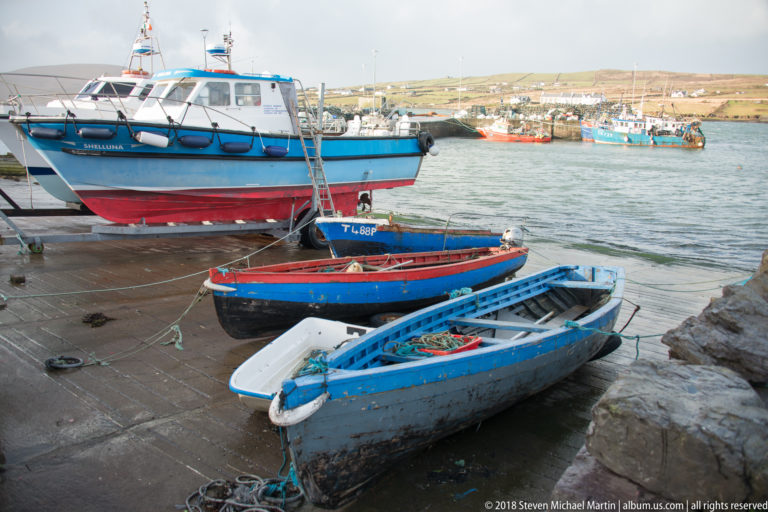 Three small open boats lie on a concrete slipway below a larger hauled fishing vessel, with harbor boats beyond.