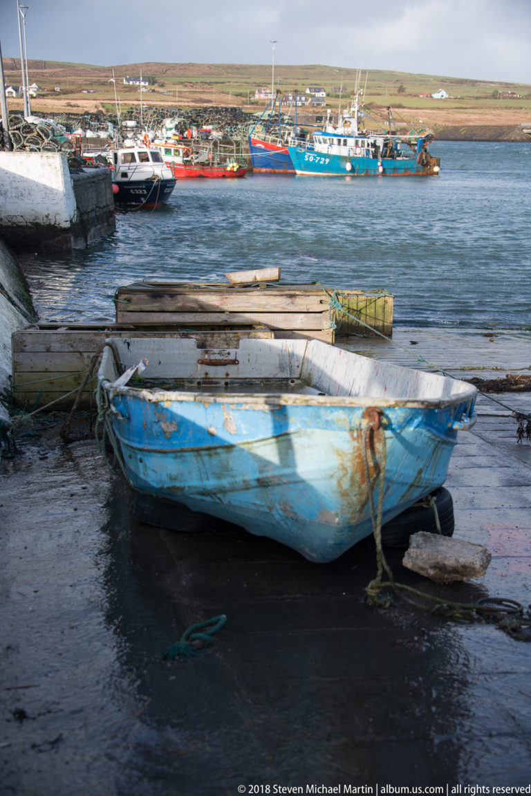 Weathered blue dinghy tied beside a wet quay, with boards and fishing gear nearby and workboats across the harbor.