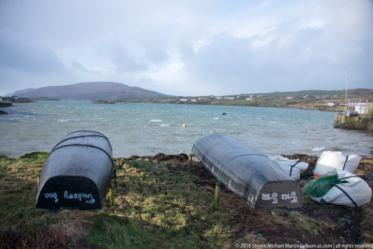Two upturned small boats resting on rough grass beside an inlet, with low hills and shoreline buildings beyond.