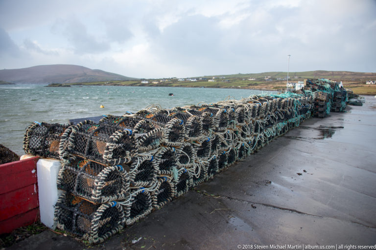 Long stacks of rope-covered fishing pots line a wet quay beside blue water, with boats and low hills beyond.