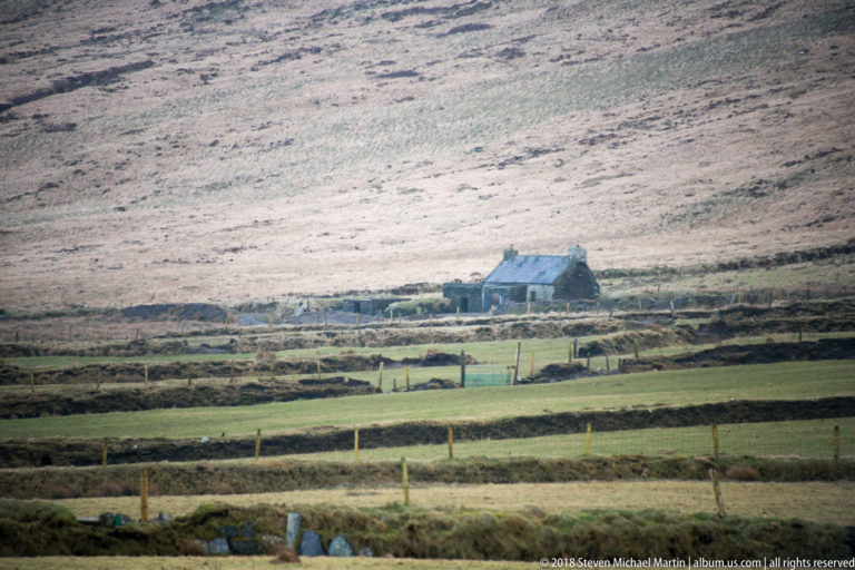 Isolated house with blue-gray roof among fenced and walled fields at the base of a broad brown hillside.