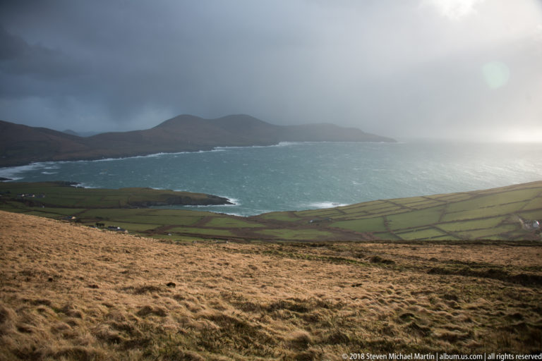 Blue-green bay seen from golden upland grass, with patchwork fields, dark mountains, and rain clouds beyond.