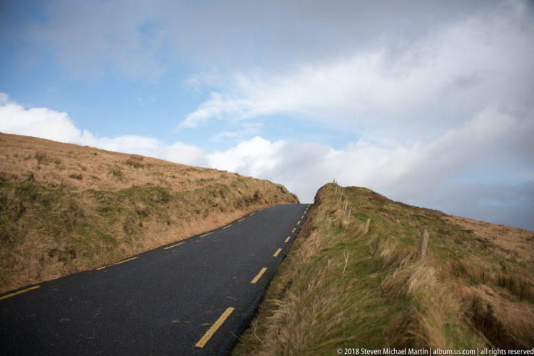 Narrow blacktop road rises to a crest between tawny grass banks beneath blue sky and broken clouds.