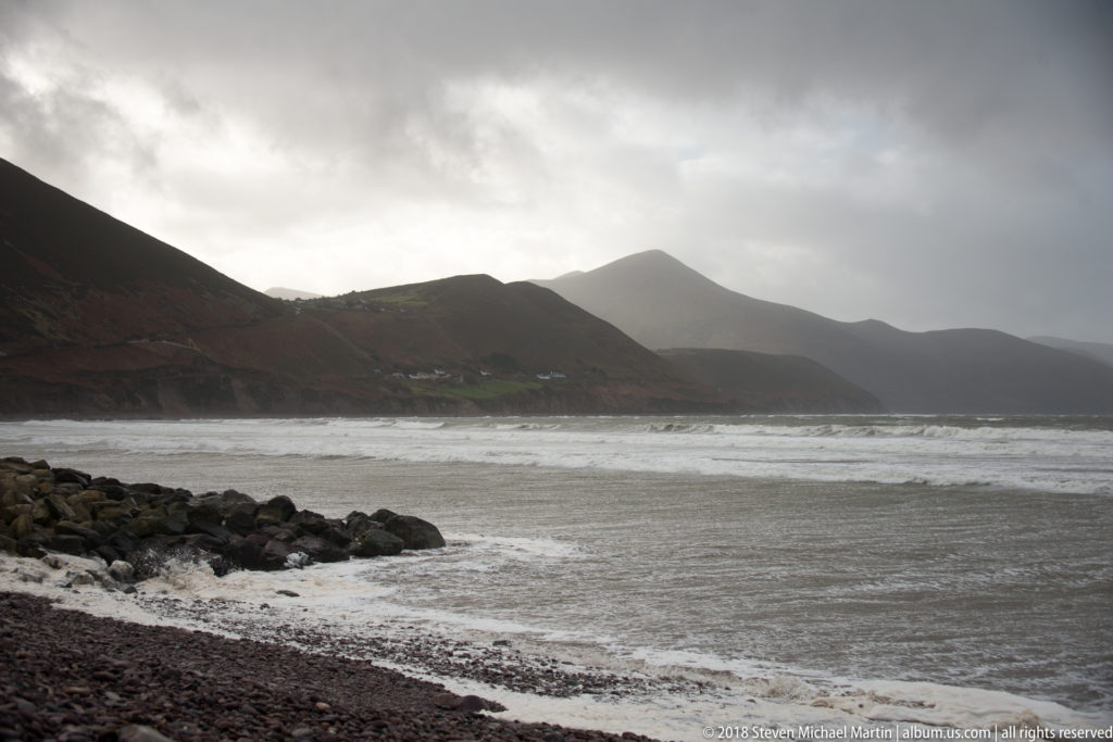 Dark mountain ridges recede beside a gray Atlantic bay, with white surf and rain clouds overhead.