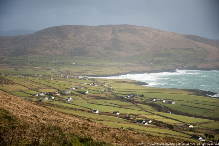 Patchwork green fields and scattered white houses slope toward a surf-filled cove below a broad brown mountain.
